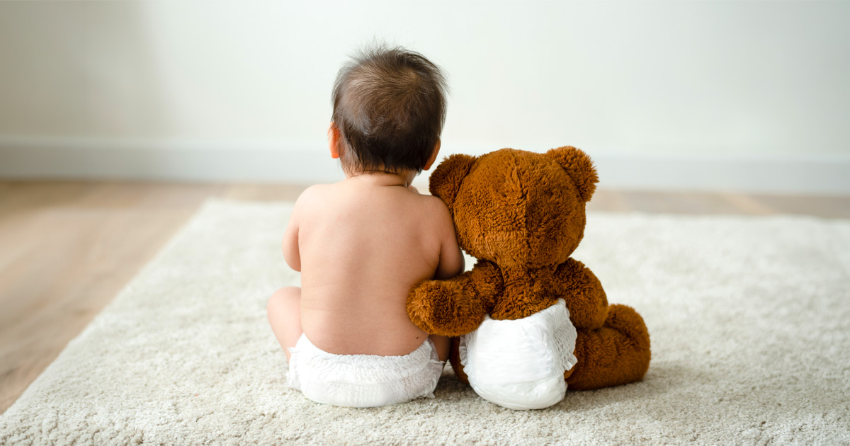 Diapered baby sitting with a stuffed teddy bear, facing away on a rug.