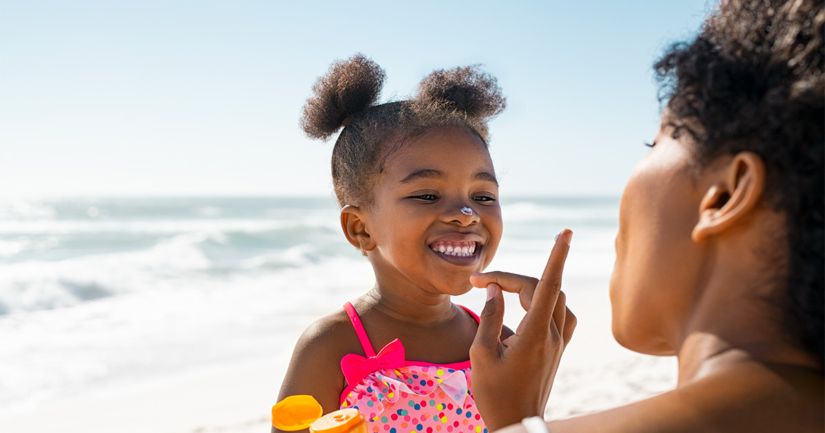 A young child at the beach having sunscreen applied to the face, with ocean waves in the background.