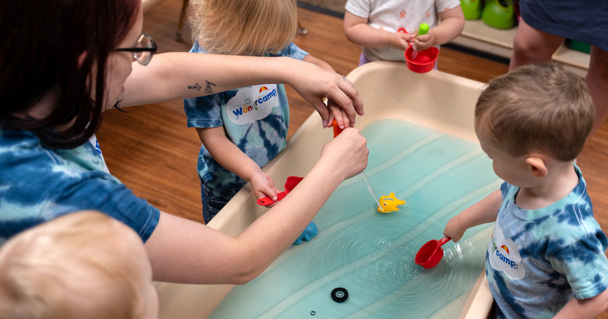A teacher and young children play with scoops and floating toys at a water sensory table.